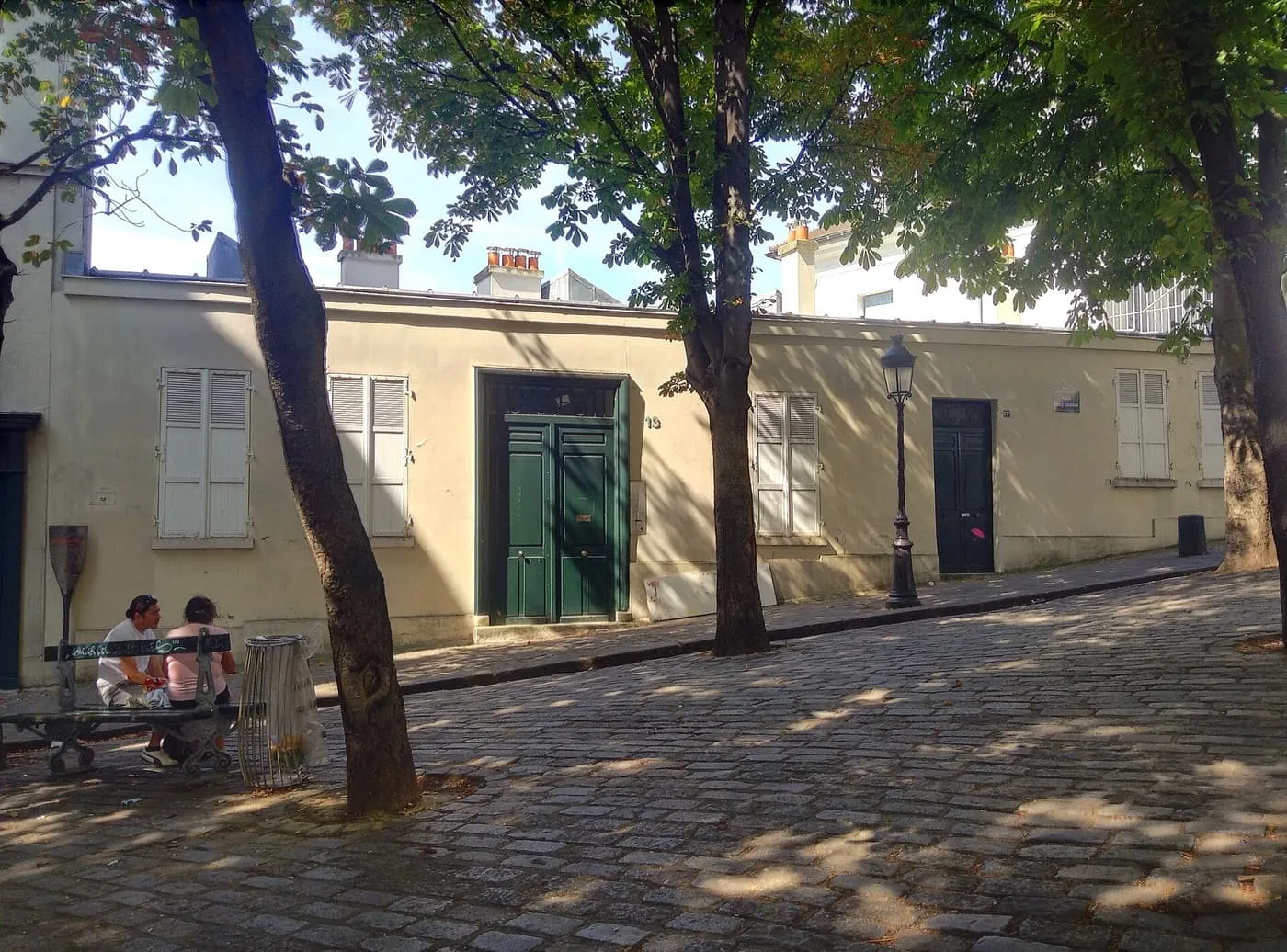 A low cream-coloured building with shuttered windows and a green double door at No. 13, on a cobbled square with chestnut trees and a bench