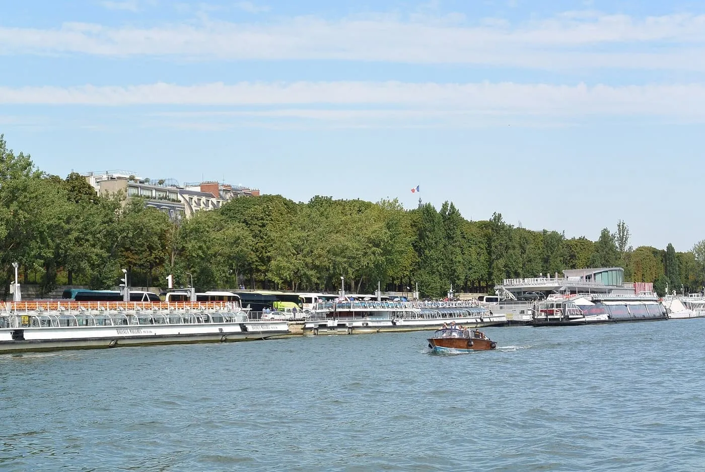 Glass-roofed Bateaux-Mouches sightseeing boats moored in a line along the Right Bank pier, with trees behind and a small wooden motorboat passing