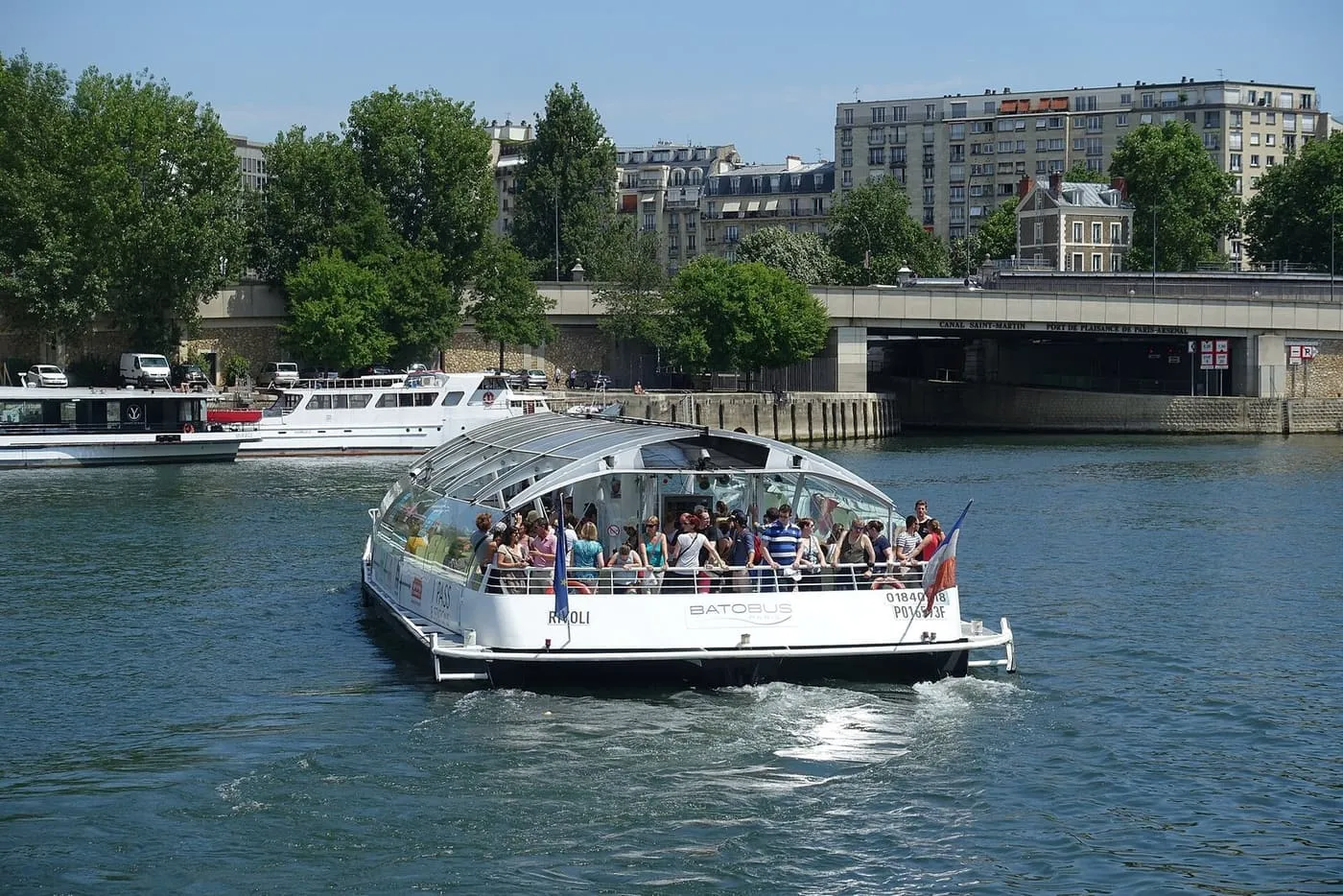 A white glass-roofed Batobus river bus with passengers on its rear deck, on the Seine beside tree-lined quays