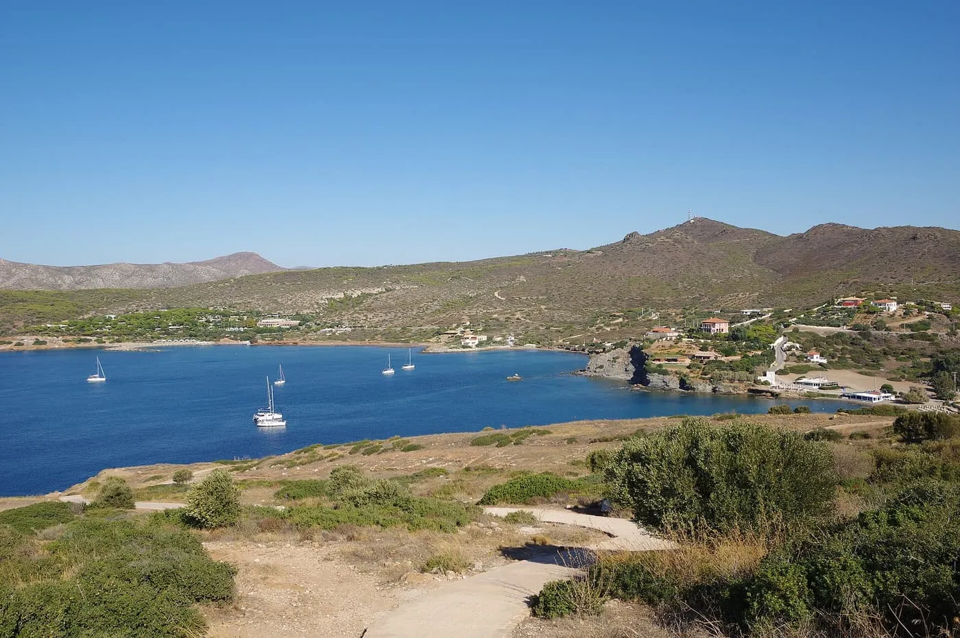 A sheltered blue bay with sailing boats at anchor, below hills with scattered houses, seen from the headland