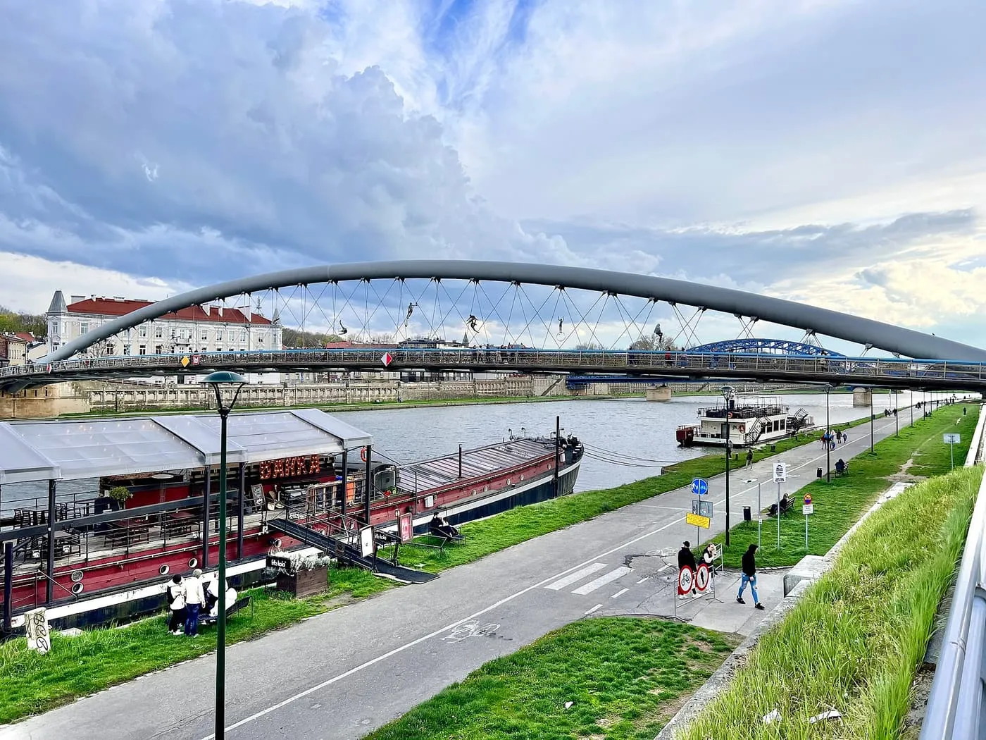 The curved steel arch of the Father Bernatek footbridge over the Vistula, with a red-hulled barge and a white river boat moored beside the embankment path