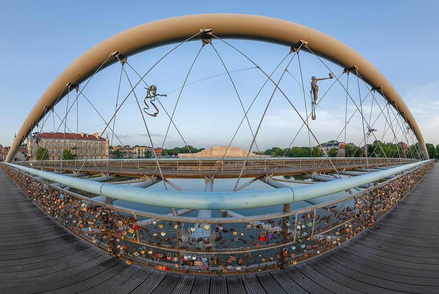 The steel arch of the Father Bernatek footbridge from the deck, with two acrobat sculptures hung from the cables and love padlocks on the railing