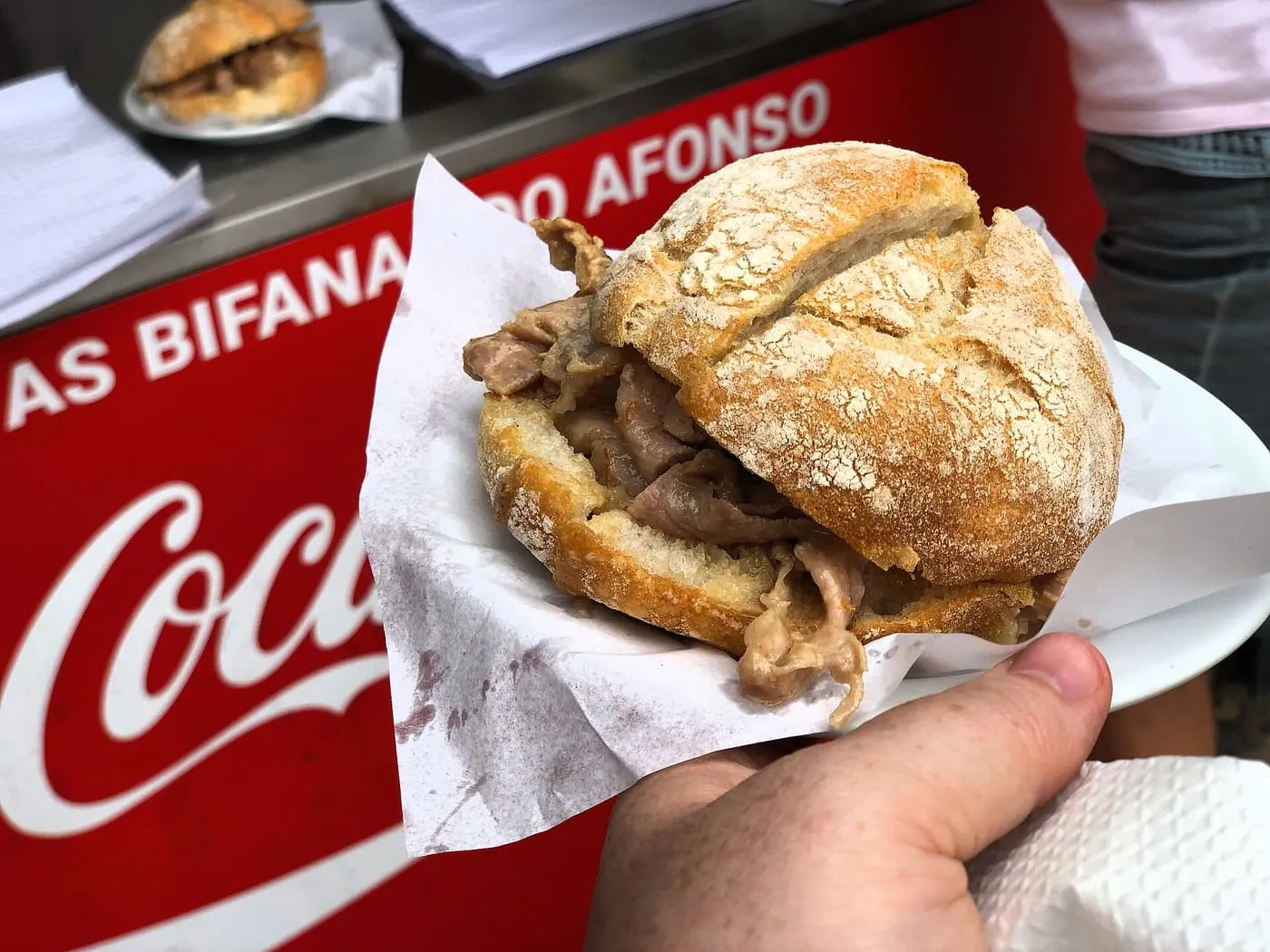 A hand holding a bifana, pork in a crusty roll on paper, in front of a red counter with the name As Bifanas do Afonso