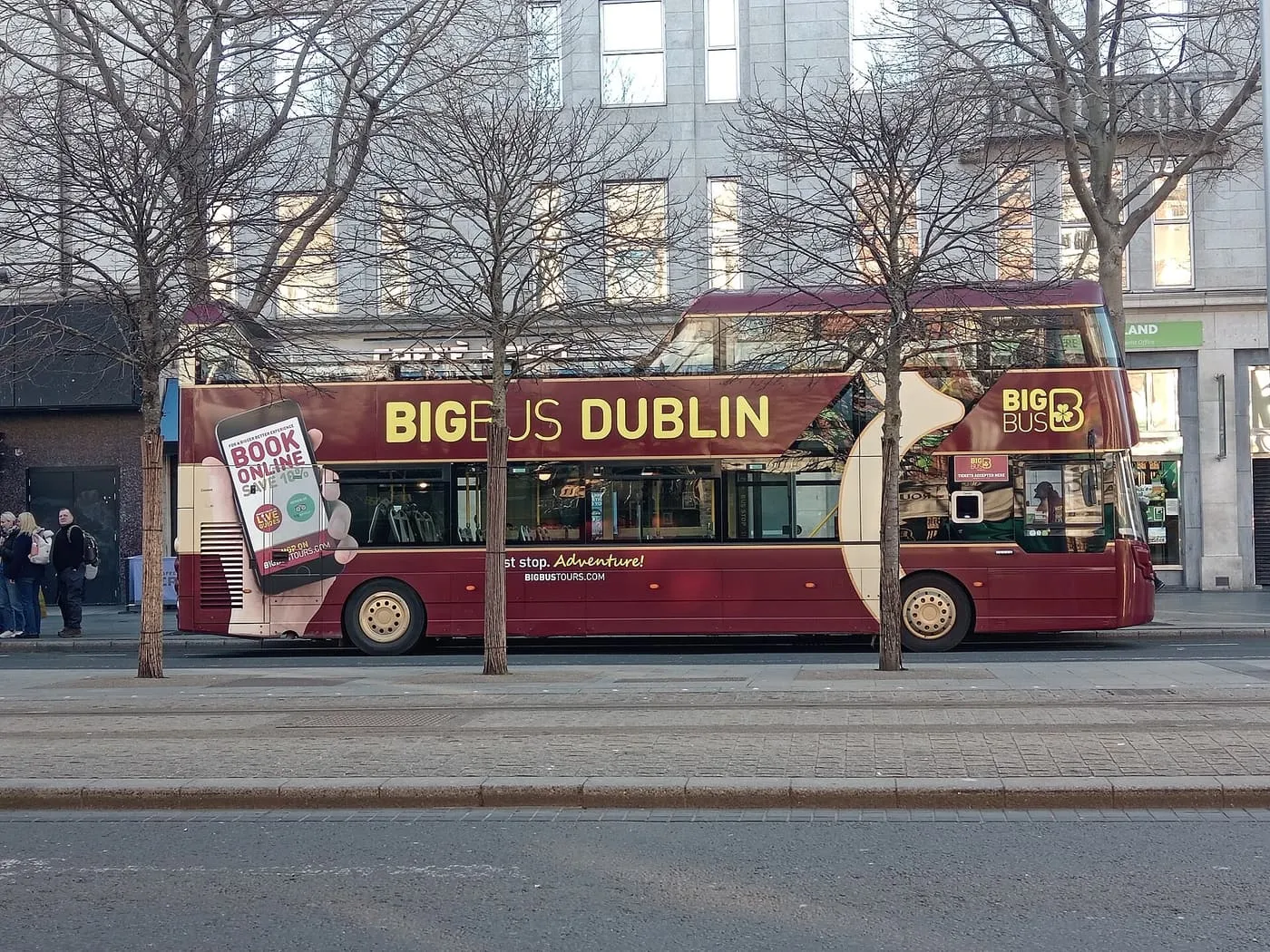 A maroon open-top double-decker with Big Bus Dublin lettering stopped at a kerb behind bare winter trees