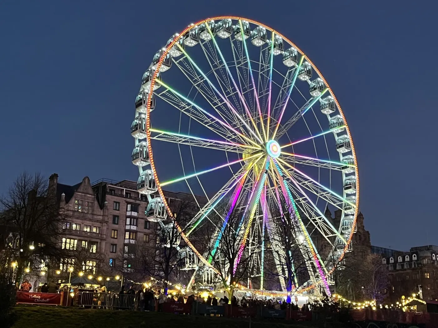 A big wheel with colour-changing spokes and a glowing rim against a dark blue sky, with bare trees and strings of market lights below