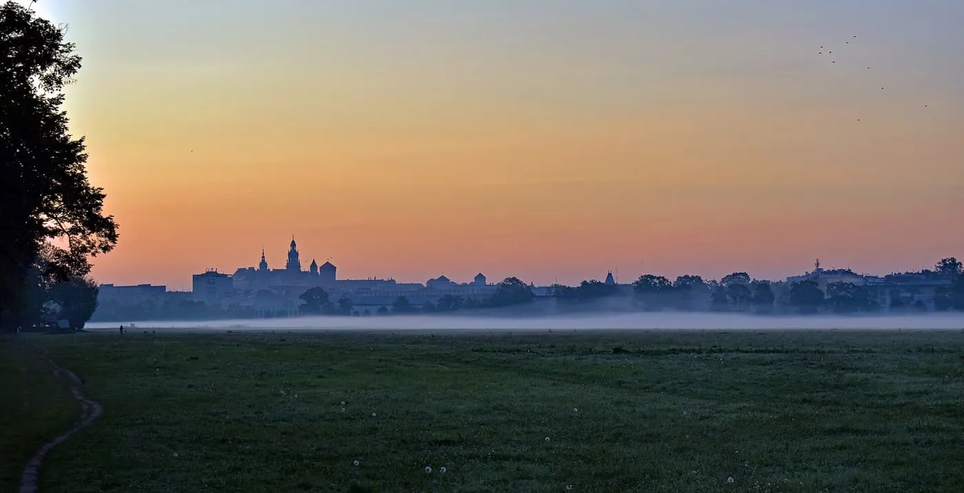 Mist lying over the Błonia meadow at dawn, with the silhouette of the Wawel on the skyline under an orange sky