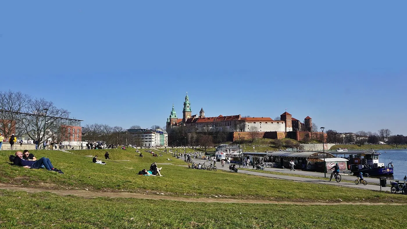 Czerwieński Boulevard on the Vistula, with boats moored along the embankment below the red roofs of the Wawel and people sitting on the grass slope