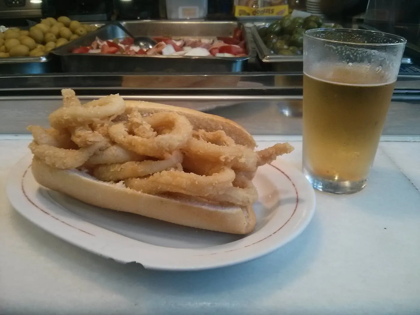 A bread roll piled with fried squid rings on a plate on a bar counter, beside a glass of beer