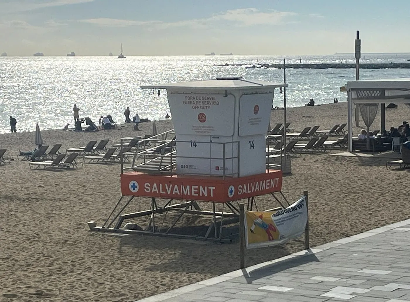 A white lifeguard tower on the sand of Bogatell beach, marked off duty with the 112 emergency number, with sun loungers and the sea behind