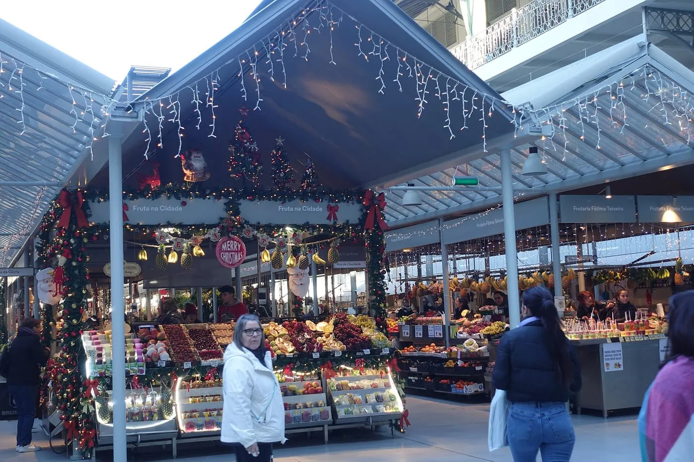 A fruit stall dressed in Christmas garlands and lights under the glass roof of the Mercado do Bolhão, with shoppers in front
