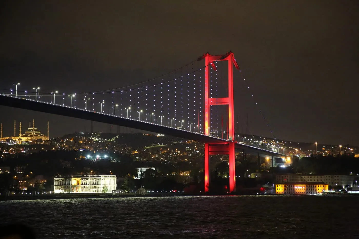 The Bosphorus Bridge lit at night, its tower glowing red and its cables studded with white lights, with a floodlit palace on the shore below