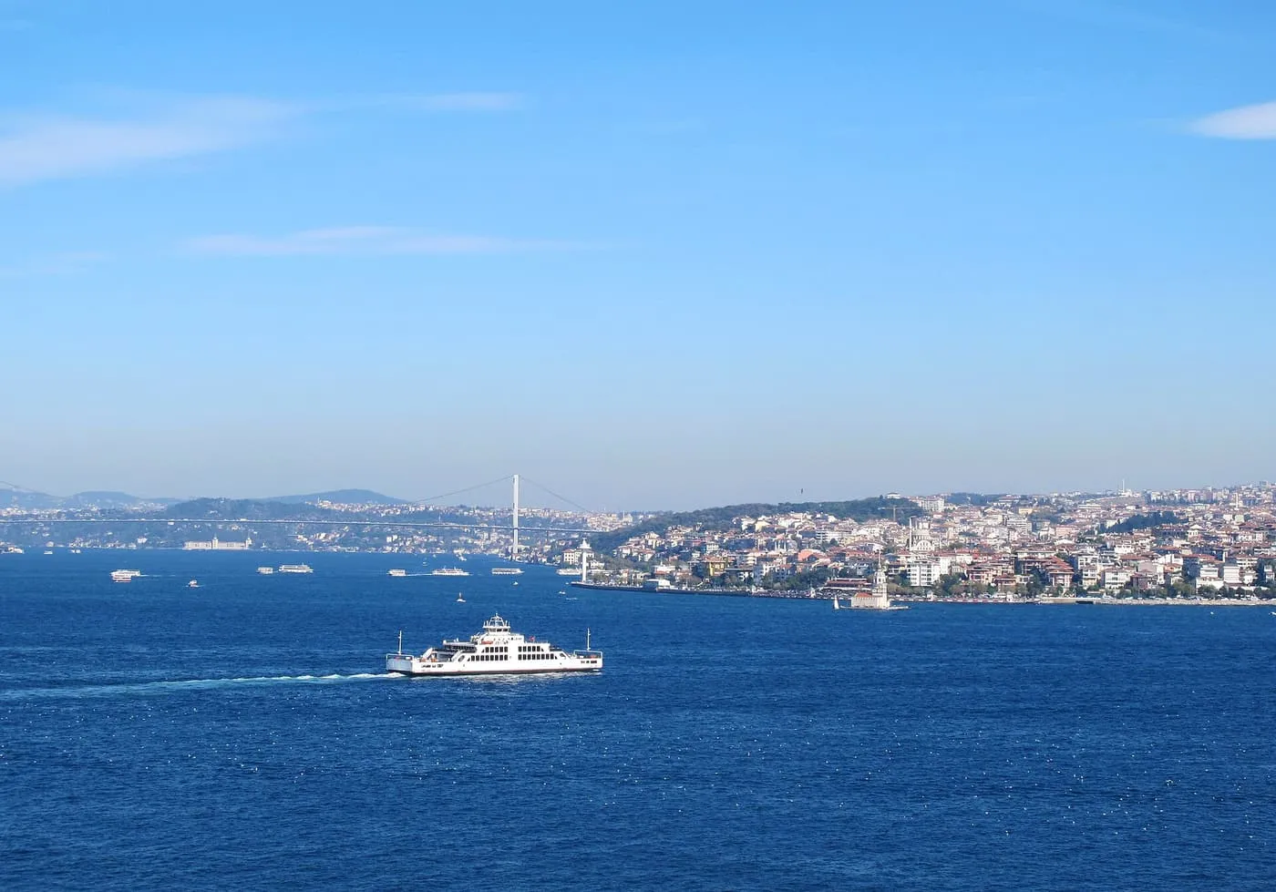 A white car ferry crossing the deep blue Bosphorus under a clear sky, with the Asian shore and a bridge in the distance