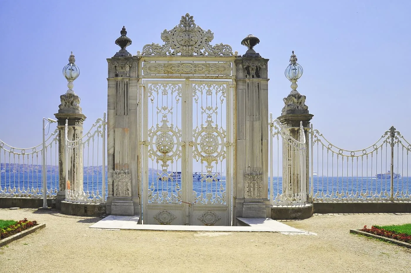 A tall white wrought-iron gate between stone piers on the Bosphorus shore, with the strait and ships behind it