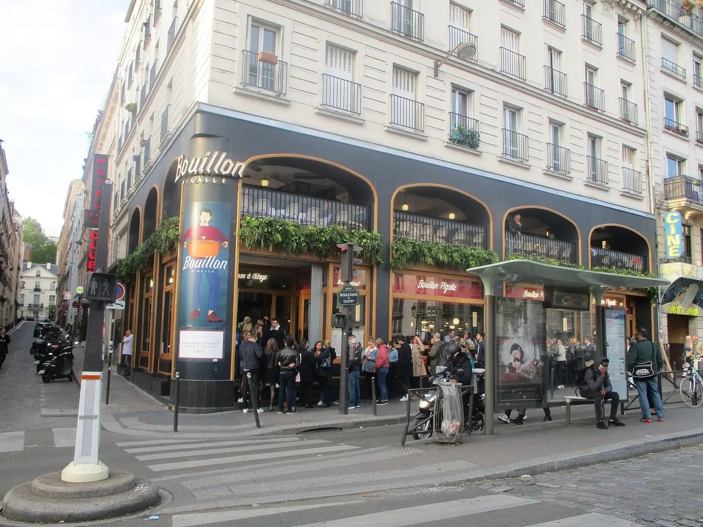 A queue of people on the pavement outside the dark glass-and-wood corner front of Bouillon Pigalle, with its first-floor arches and hanging plants