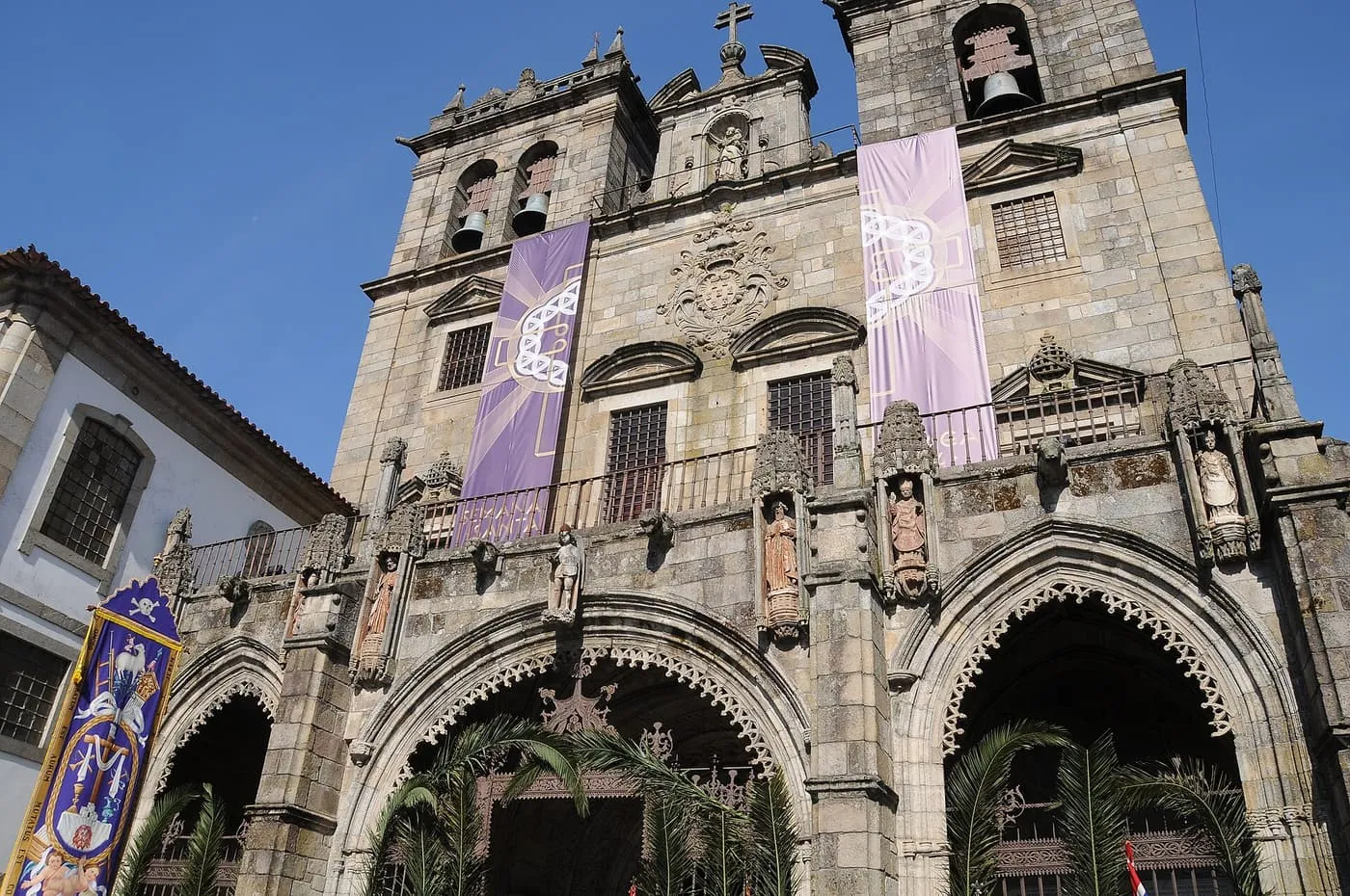 The carved granite west front of Braga Cathedral, with two bell towers and pointed arches, hung with purple banners