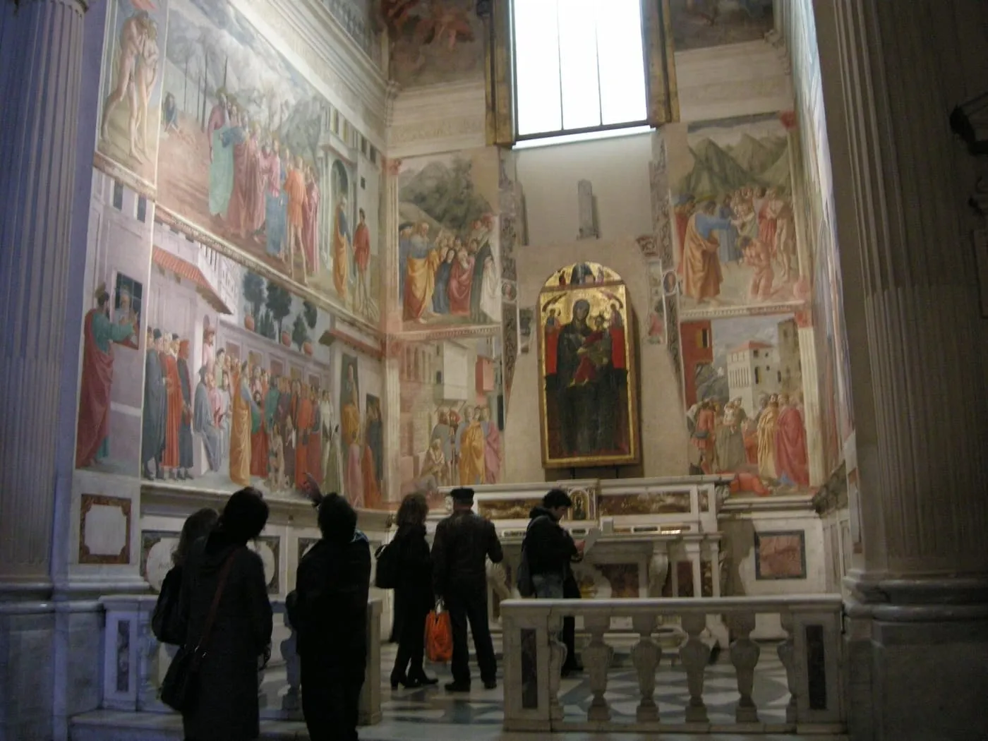 Visitors at the rail of the Brancacci Chapel, with frescoes on the side walls and a painted Madonna above the altar