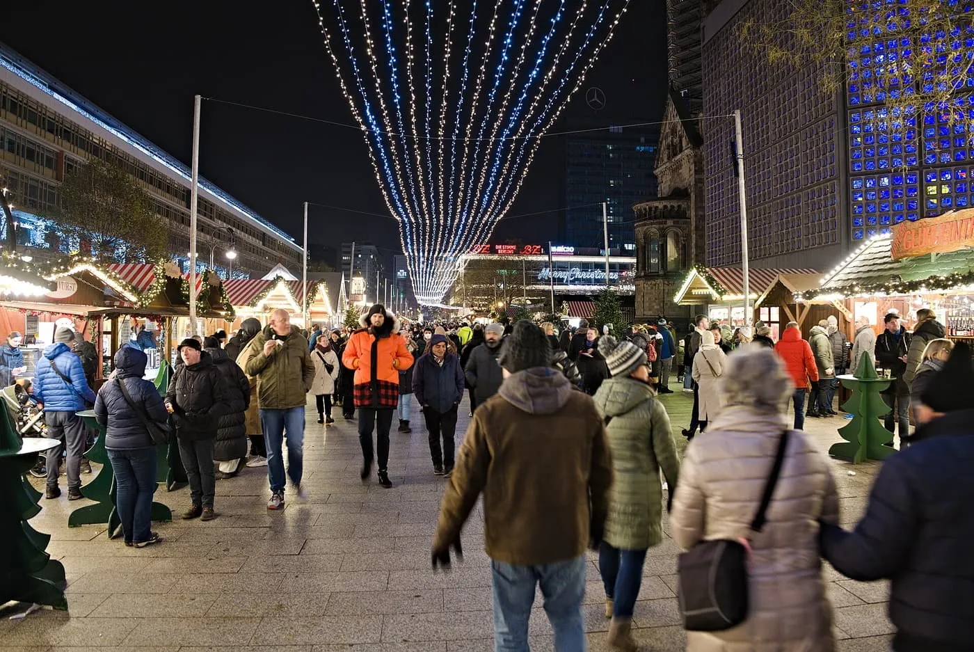 Visitors walking between market stalls beneath strings of lights beside the blue glass wall of the Kaiser Wilhelm Memorial Church