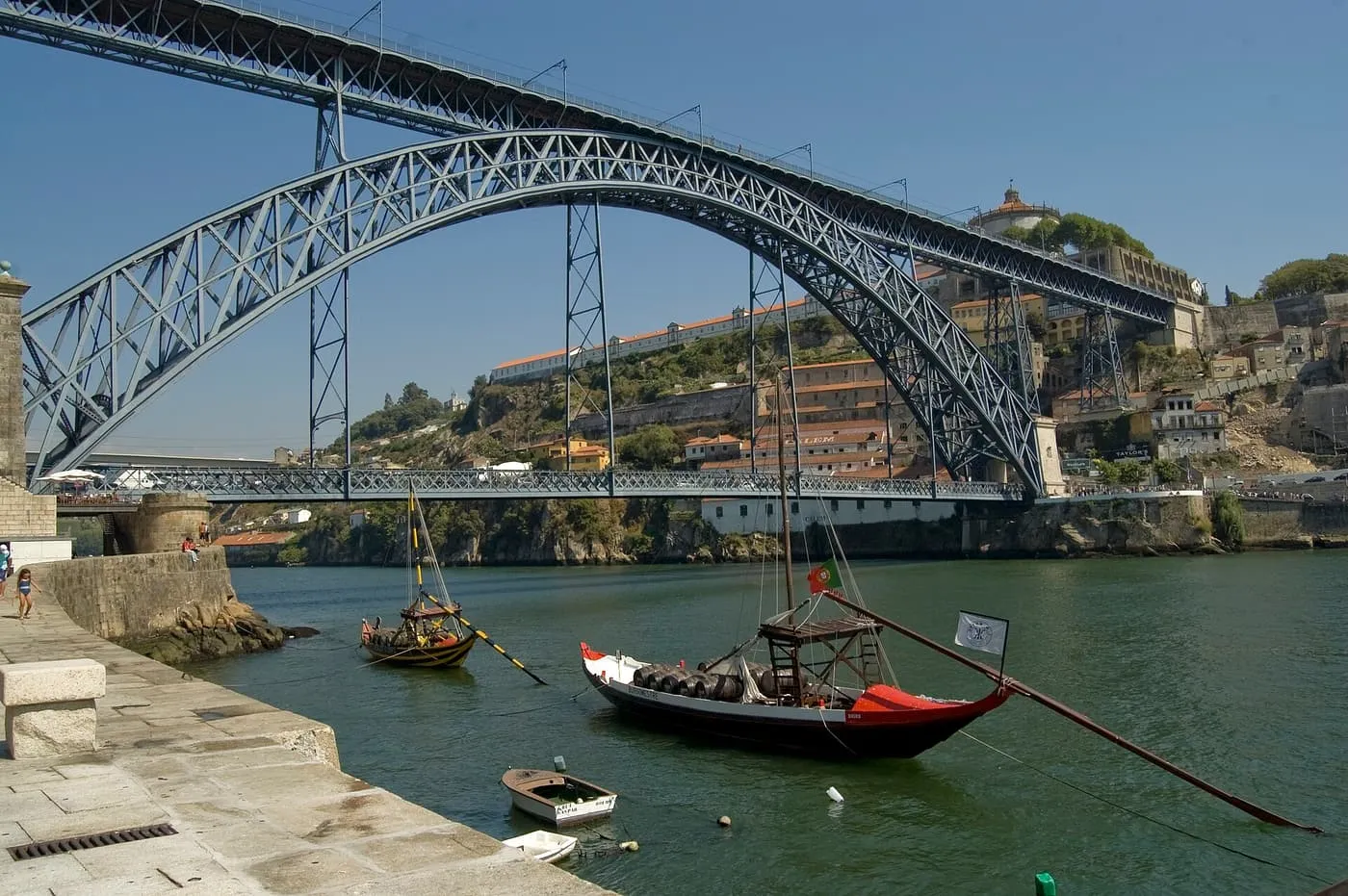 The two iron arches of the Dom Luís I bridge over the Douro seen from the Ribeira quay, with rabelo boats moored in the foreground