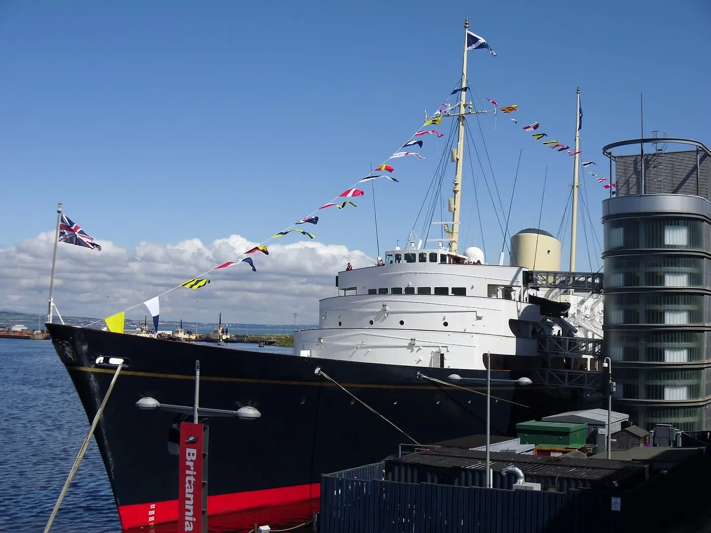 The dark-hulled Royal Yacht Britannia dressed with flags beside the glass tower of Ocean Terminal in Leith