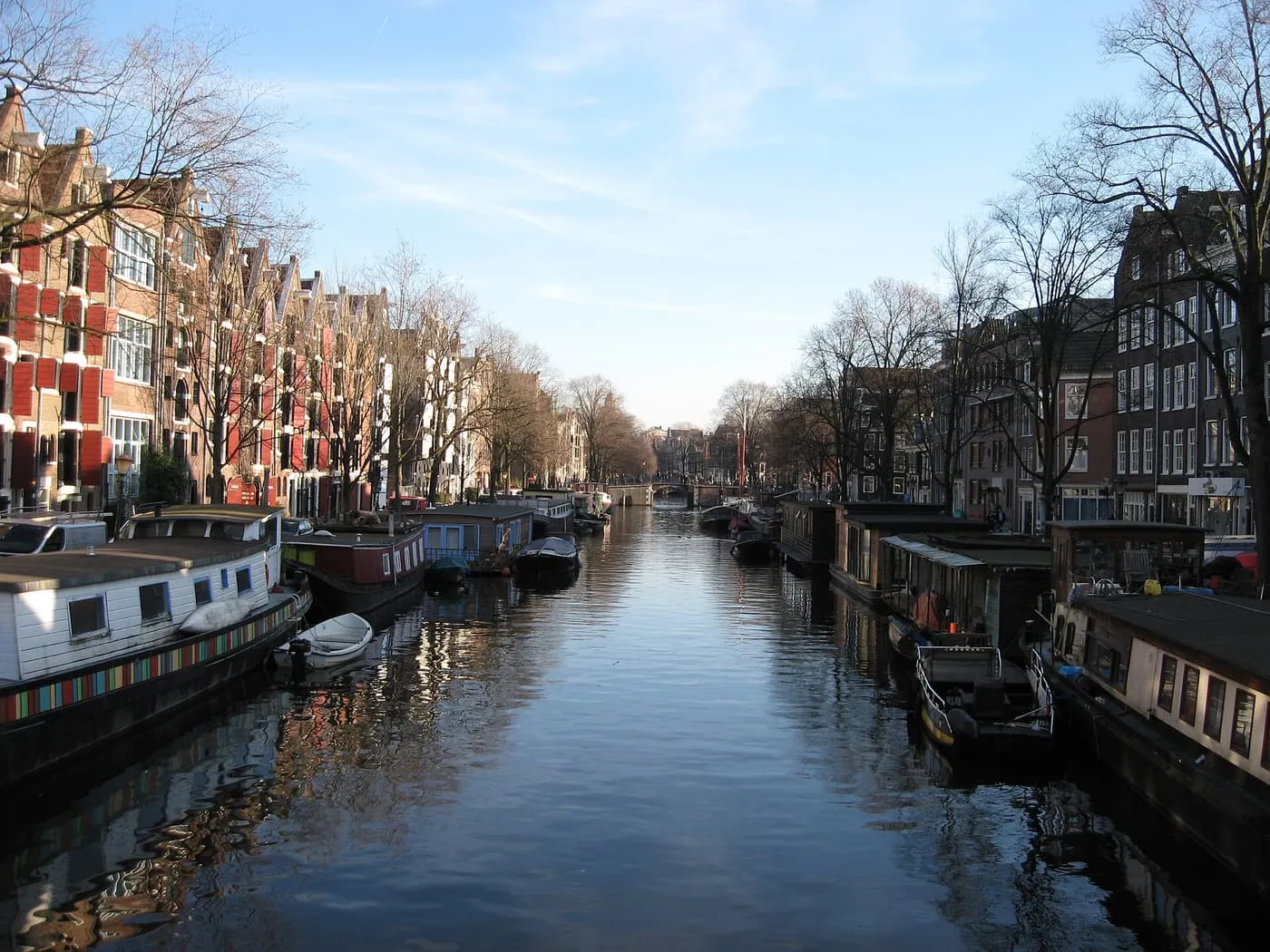 A wide canal lined with houseboats, with gabled warehouses with red shutters on the left and a bridge at the far end
