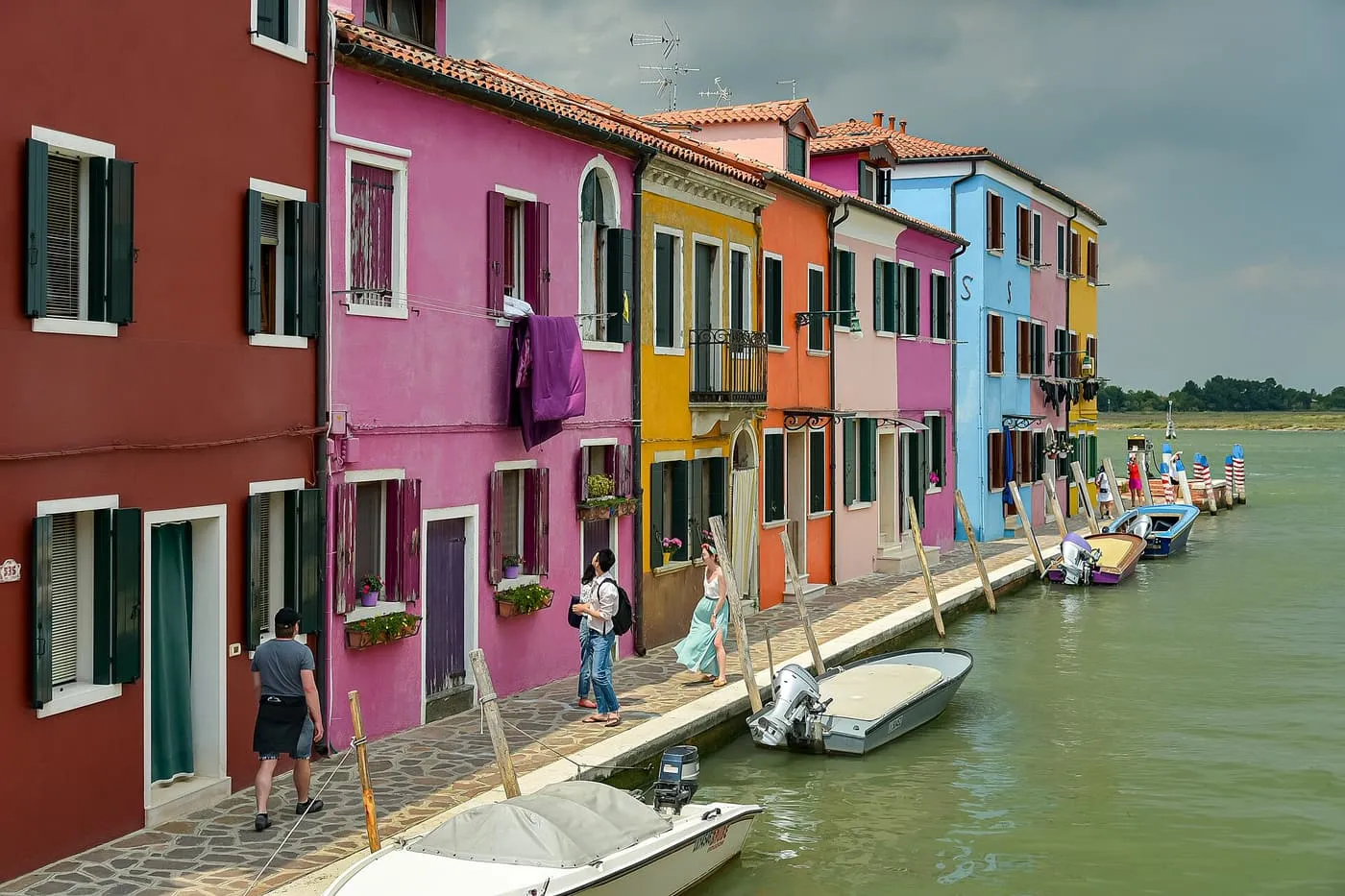 A row of brightly painted houses beside a canal on Burano, with small boats moored along the quay and the lagoon beyond