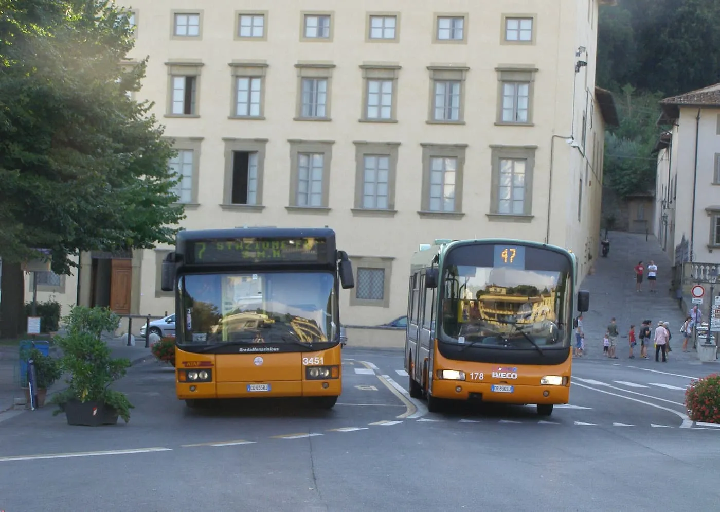 Two orange city buses in a square in Fiesole, with a tall pale building behind them