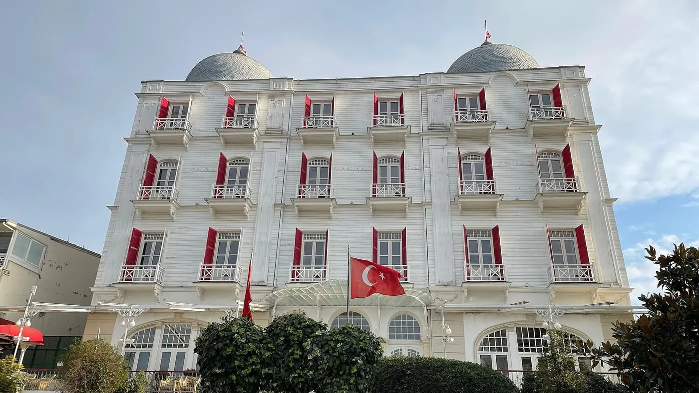 The white wooden façade of the Splendid Palace hotel on Büyükada, with red shutters, two grey domes and a Turkish flag