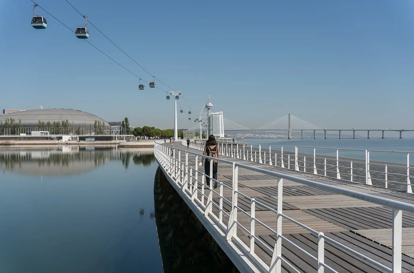 Cable cars hanging above a wooden boardwalk beside the Tagus, with the Vasco da Gama Bridge on the horizon