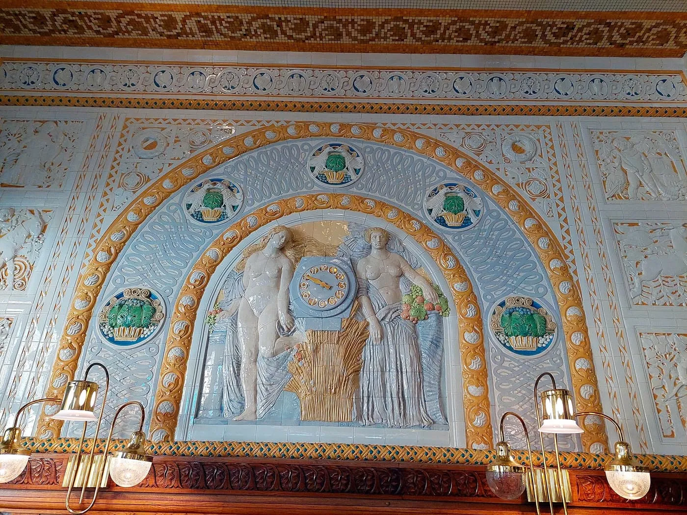 A glazed ceramic wall panel in Café Imperial showing two figures either side of a clock, framed by an arch of blue and ochre tiles