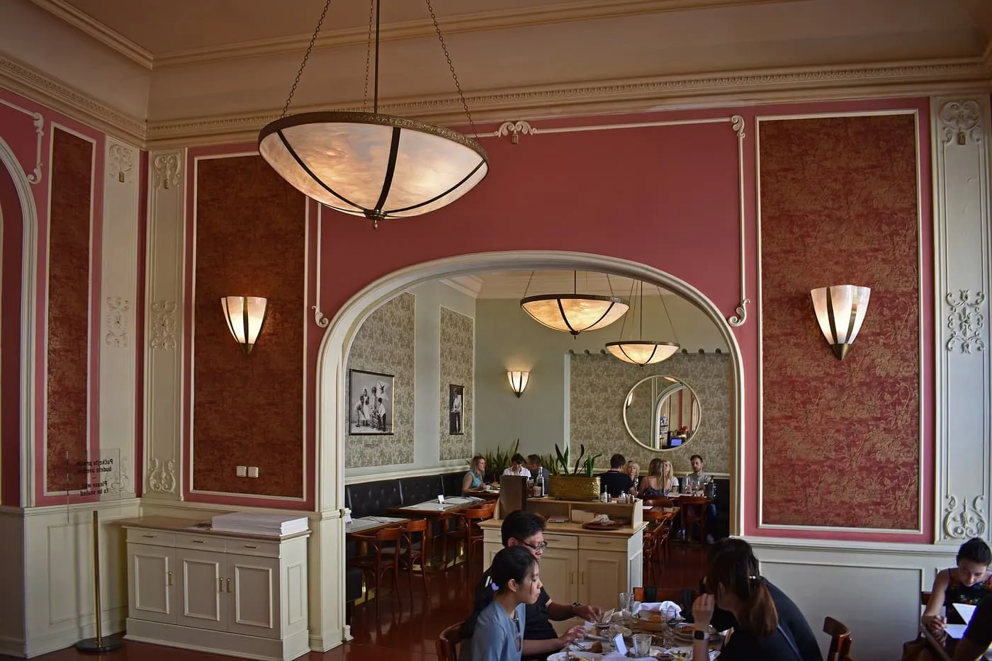 A high-ceilinged dining room at Café Louvre with pink walls, cream mouldings, shell-shaped pendant lamps and an arch opening onto a second room