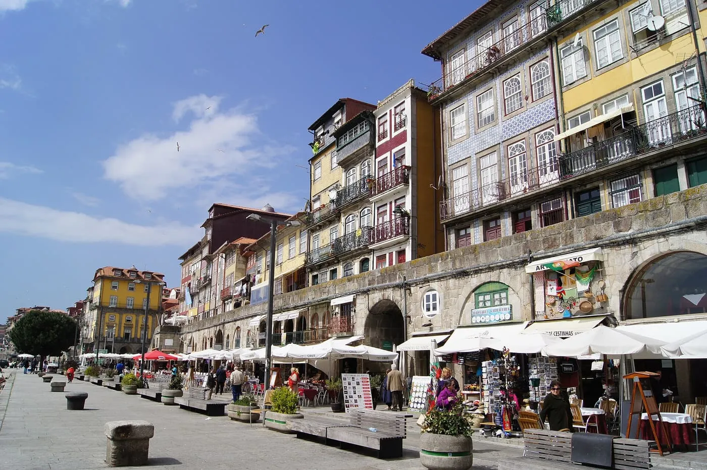 A waterfront street running along arcaded houses, with café terraces under white parasols and a few people walking
