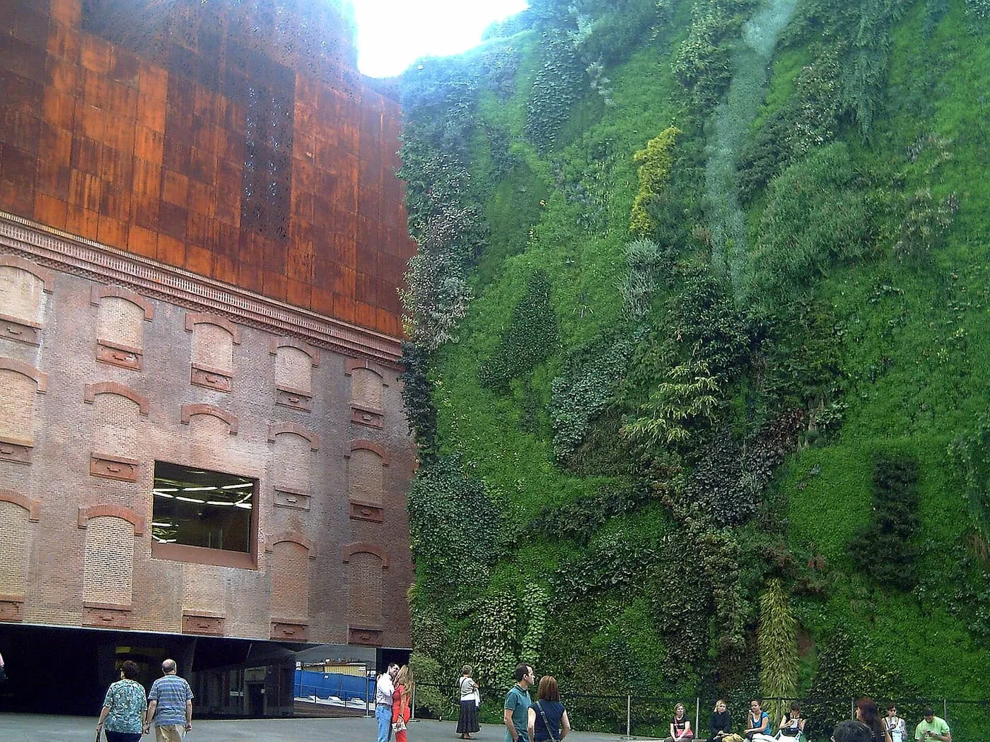 The brick and rust-coloured walls of CaixaForum beside a tall wall of plants, with visitors in the square below