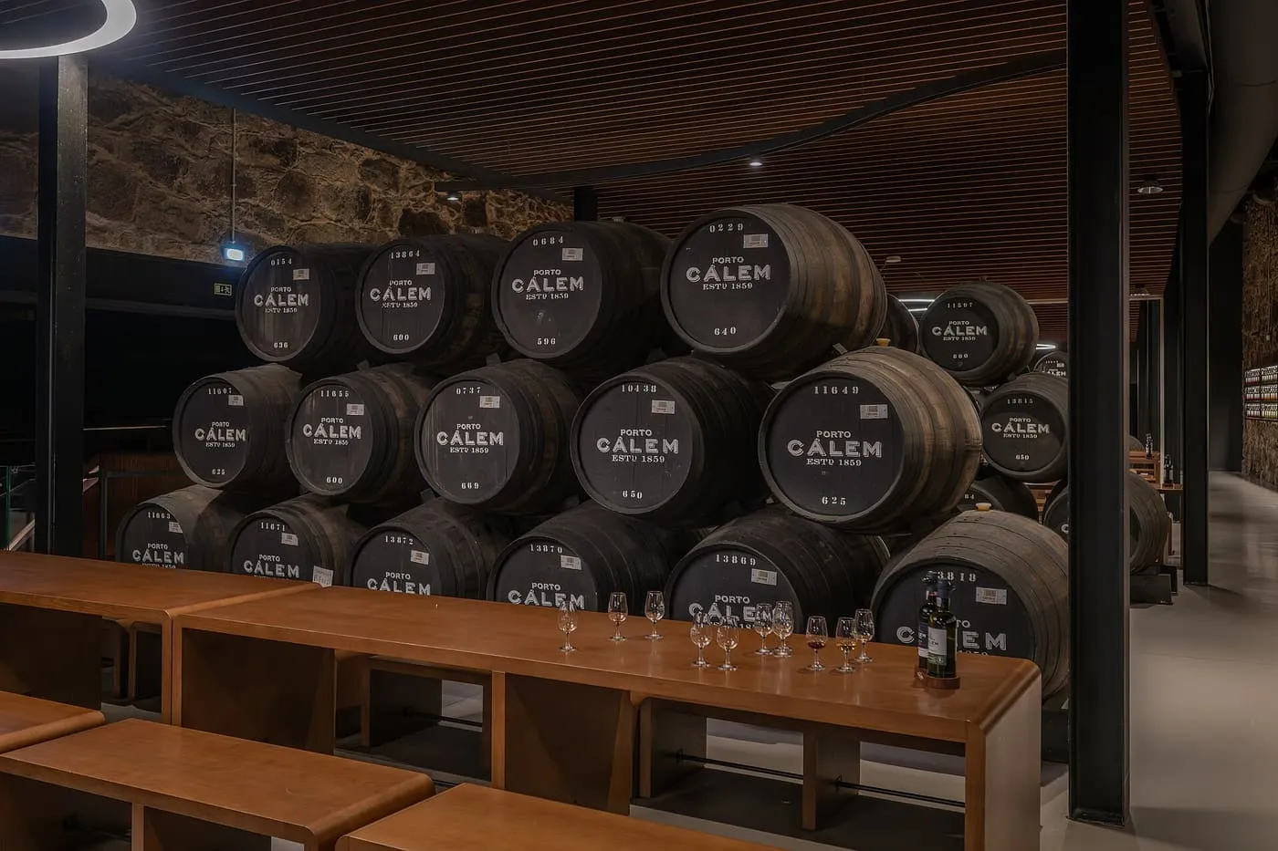 Stacked Cálem port casks in a stone-walled cellar, with a long wooden tasting table and a row of port glasses in front