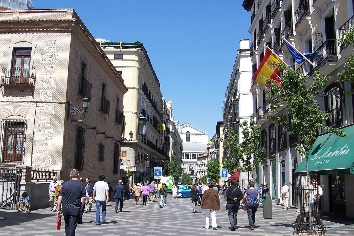 Shoppers walking along Calle del Arenal on a sunny day, between a brick palace and white apartment buildings with a Spanish flag on the right