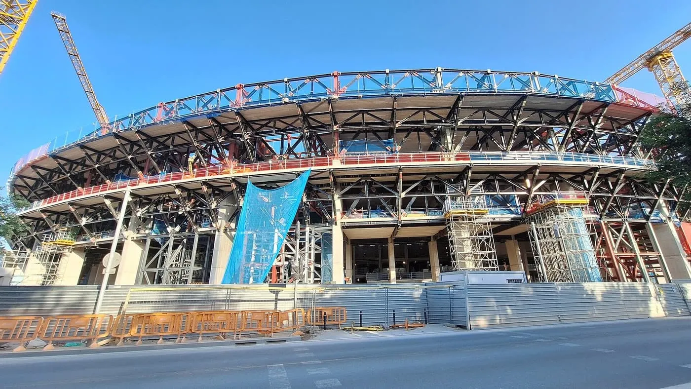 The curved steel skeleton of the stadium's upper tiers behind a street-level hoarding, with orange barriers and a blue net hanging from the frame