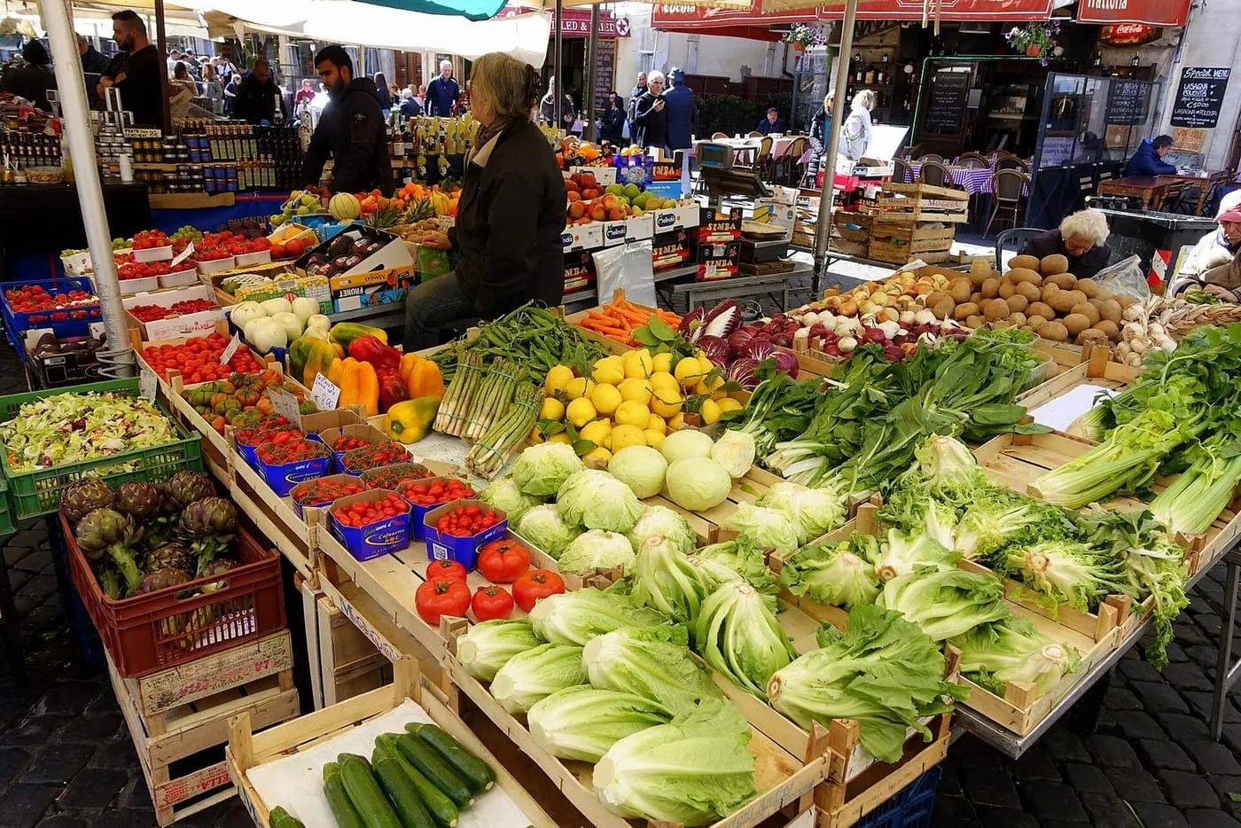 A vegetable stall at the Campo de' Fiori market, with artichokes, tomatoes, lemons, asparagus and chicory in wooden crates and café tables behind