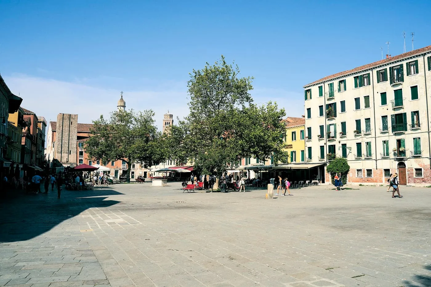 A wide paved square with plane trees and café awnings under a blue sky