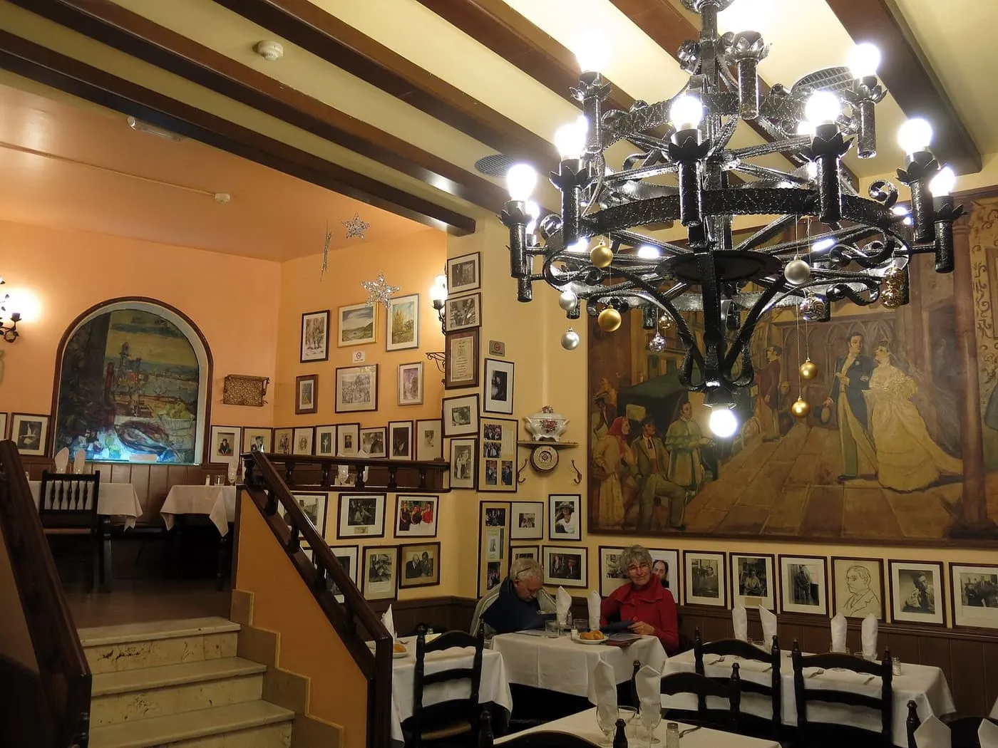 A dining room with a wrought-iron chandelier, a large painted mural of a street scene and framed photographs covering the walls