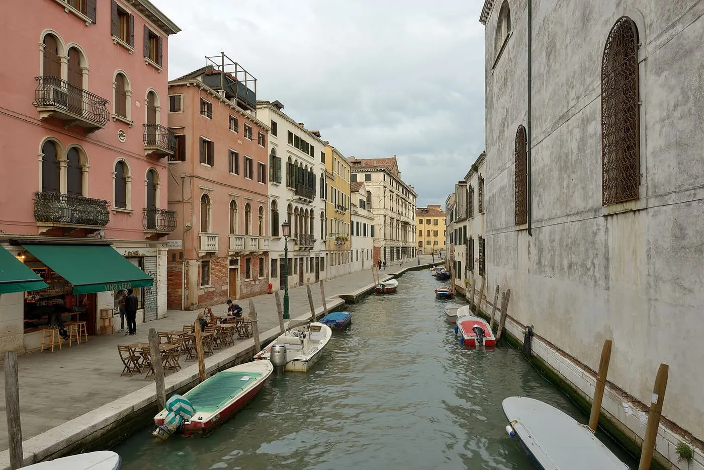 A canal in Cannaregio lined with pink and cream houses, with a wine bar's green awning on the left and boats moored along the quay