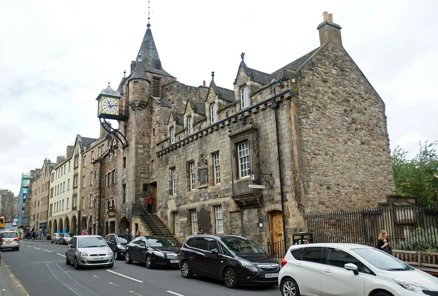 The turreted stone Canongate Tolbooth with a projecting clock, seen from the Canongate with cars parked along the kerb