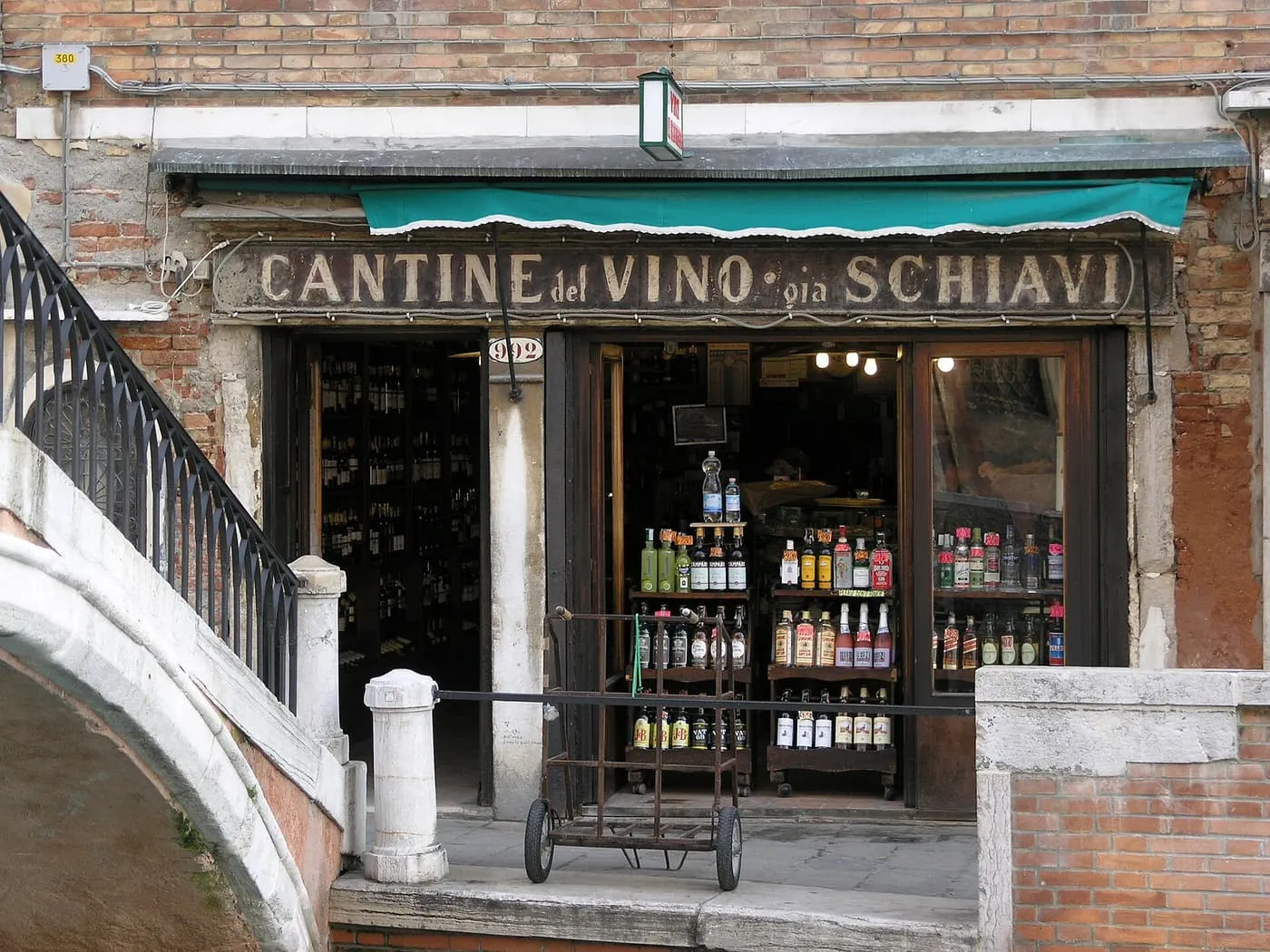 A wine shop with a painted sign reading Cantine del Vino già Schiavi, bottles on shelves inside and a bridge stair at its side
