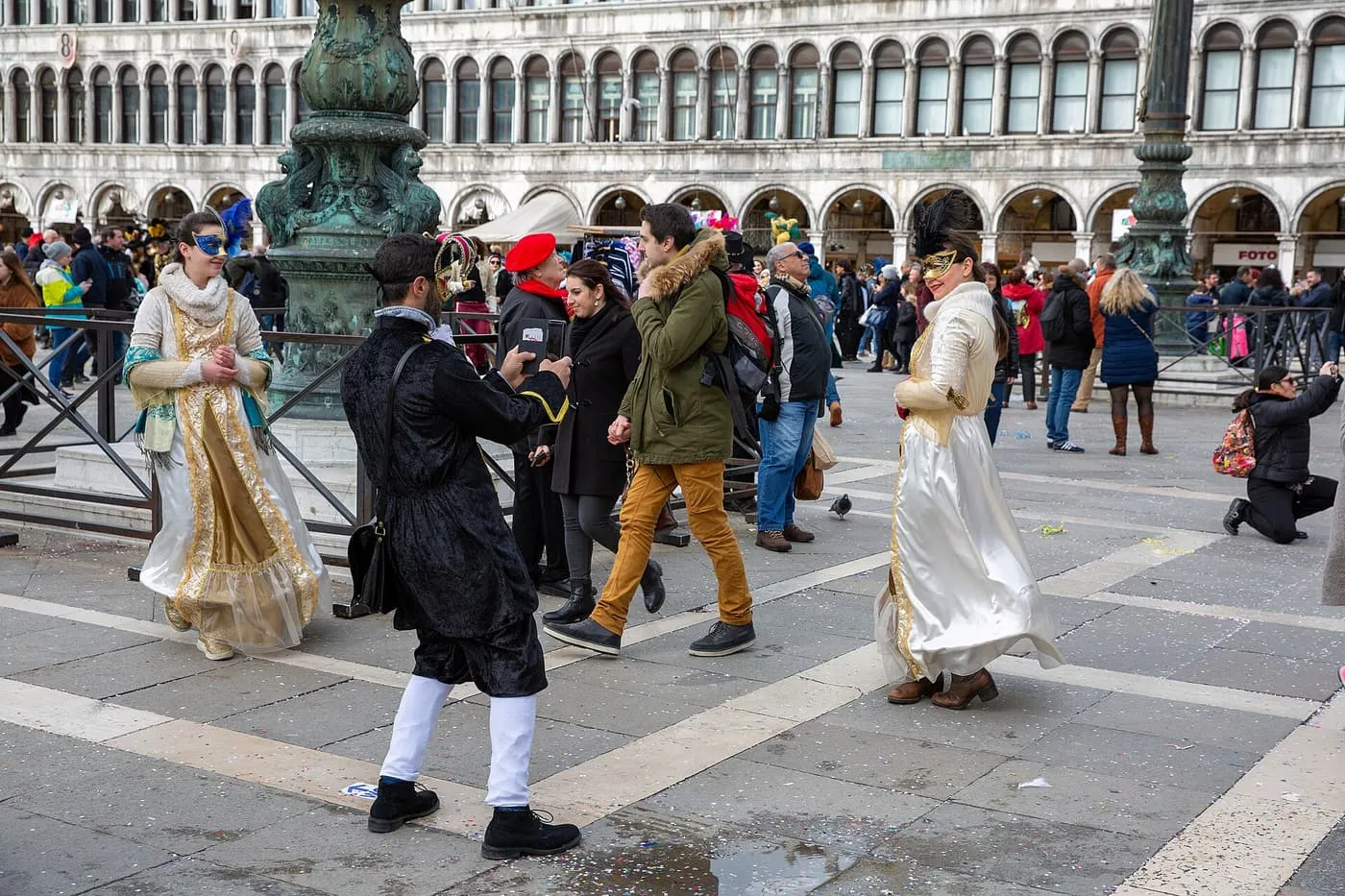 Two people in gold and white gowns and a man in a black coat and white stockings in St Mark's Square, with the arcades behind