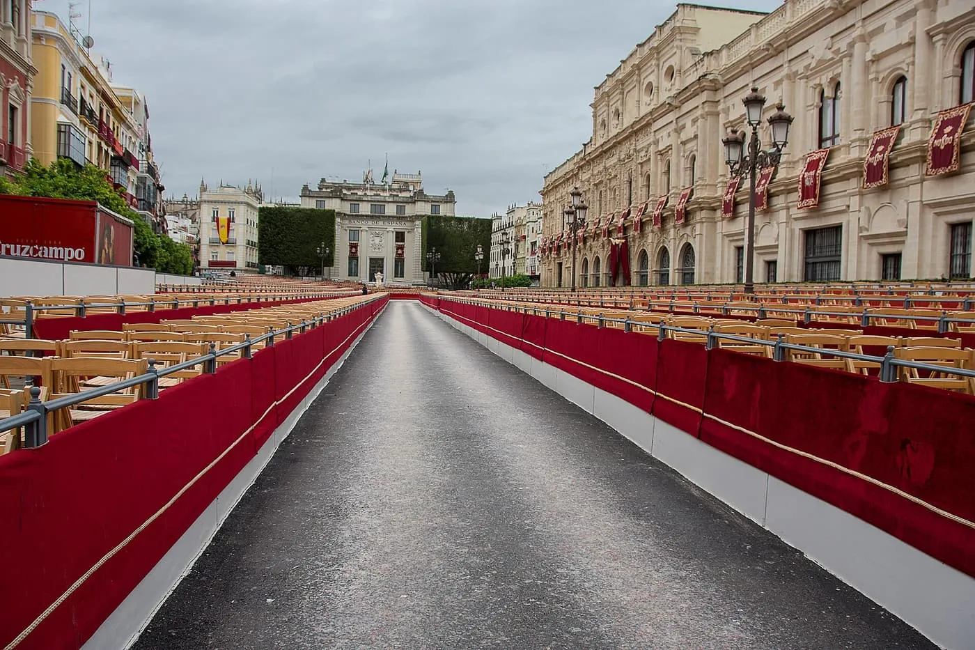 A processional route lined with rows of seats and red-draped barriers, with a stone building hung with red banners on the right