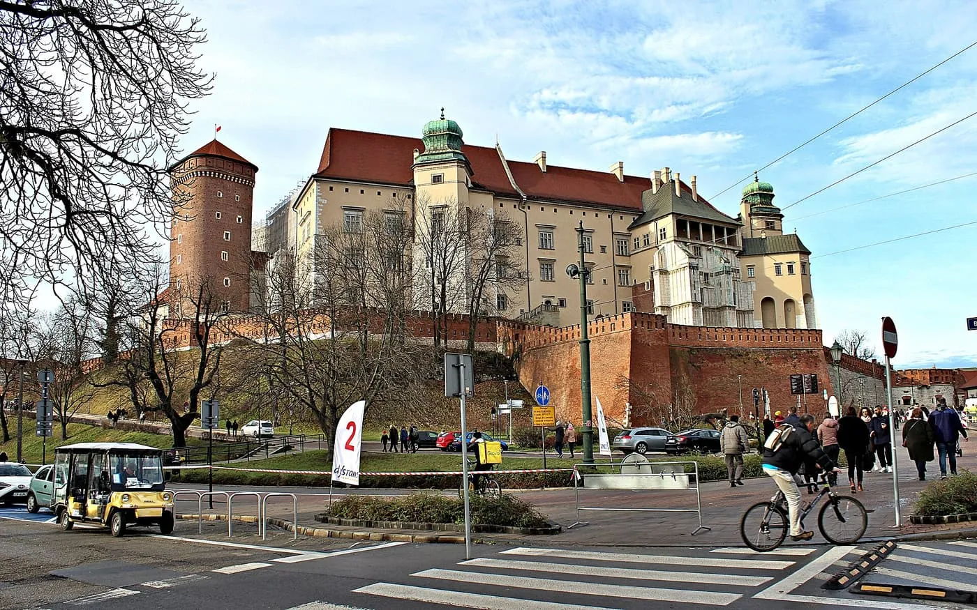 The red-brick tower and walls of the Wawel above a road crossing, with a small yellow electric cart at the lower left and a cyclist on the right