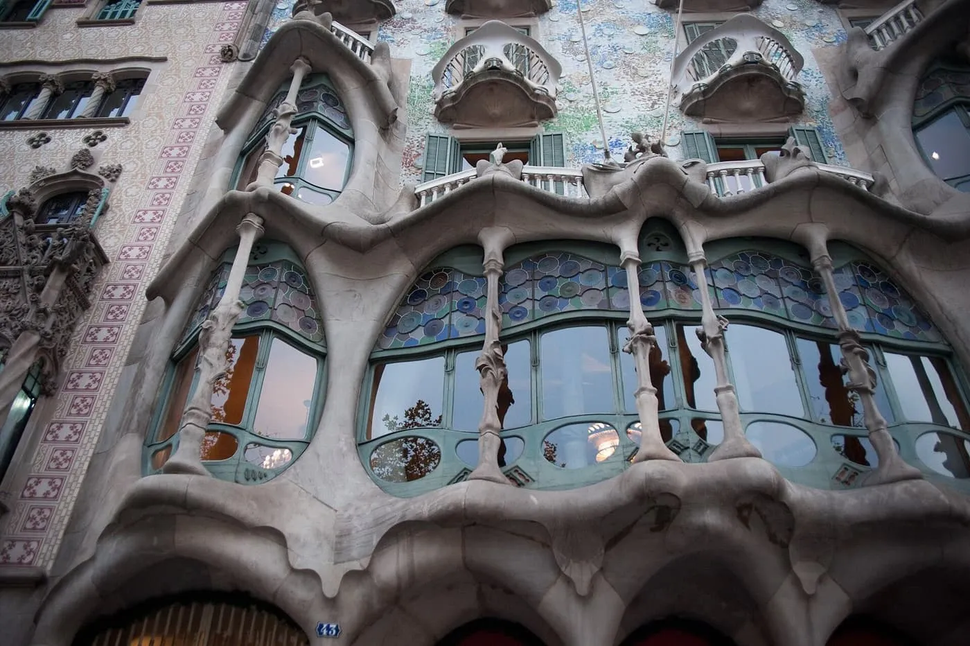 Looking up at Casa Batlló's façade: the stone-framed bay of tall windows with bone-like columns, blue glass discs above, and the mosaic wall and balconies over it