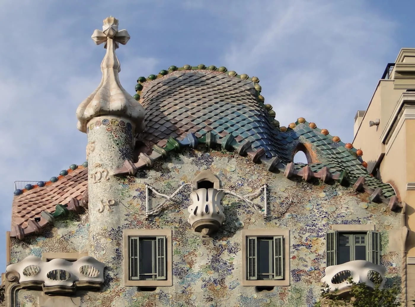 The top of Casa Batlló, a scaled roof in blue and green with a bulb-shaped tower and a four-armed cross, above a tiled front with oval windows and pale balconies