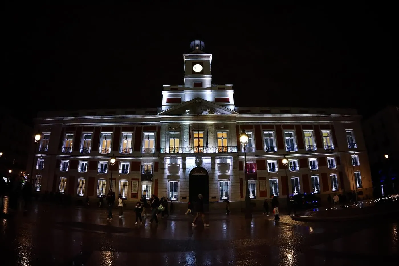 The floodlit Real Casa de Correos at night, with its clock tower, over a wet Puerta del Sol with a few people walking across