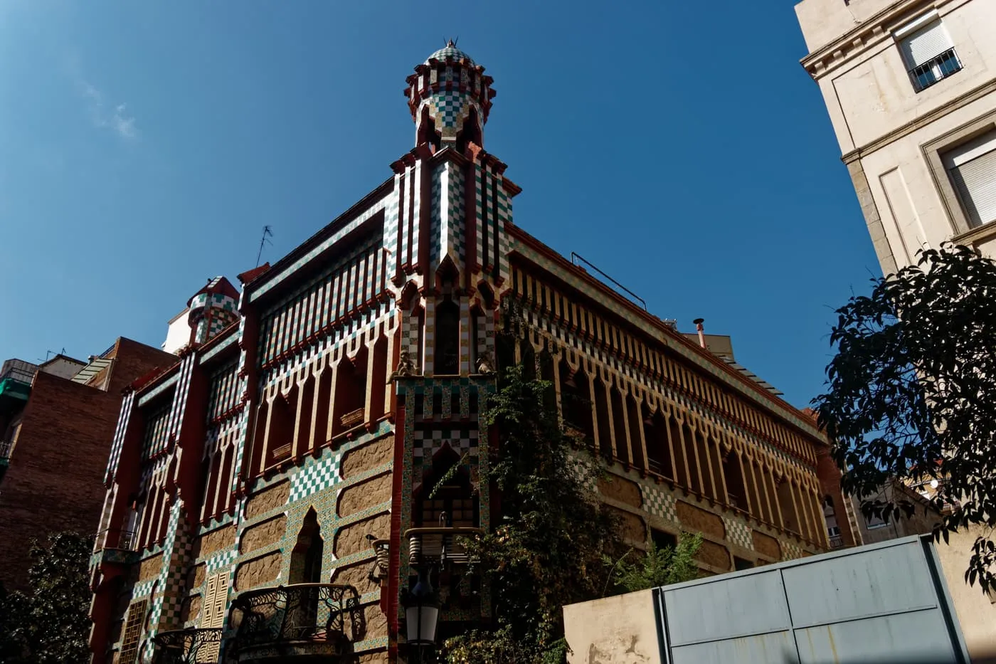 Casa Vicens from the street, a house of red brick and green and white chequered tiles with a small tower, seen from below against a blue sky