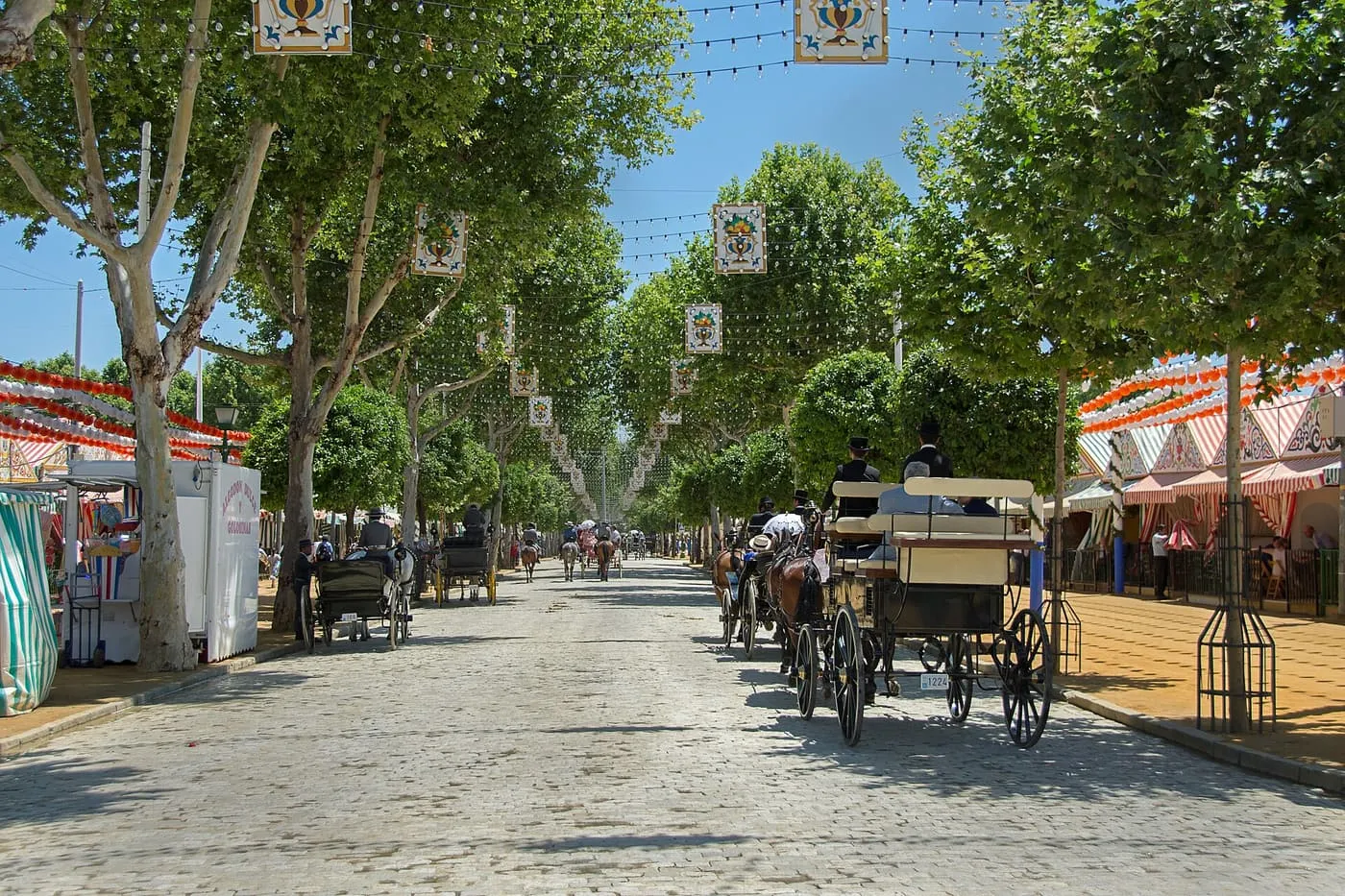 A street of striped casetas under plane trees at the Feria de Abril, with riders and a horse-drawn carriage on the cobbles