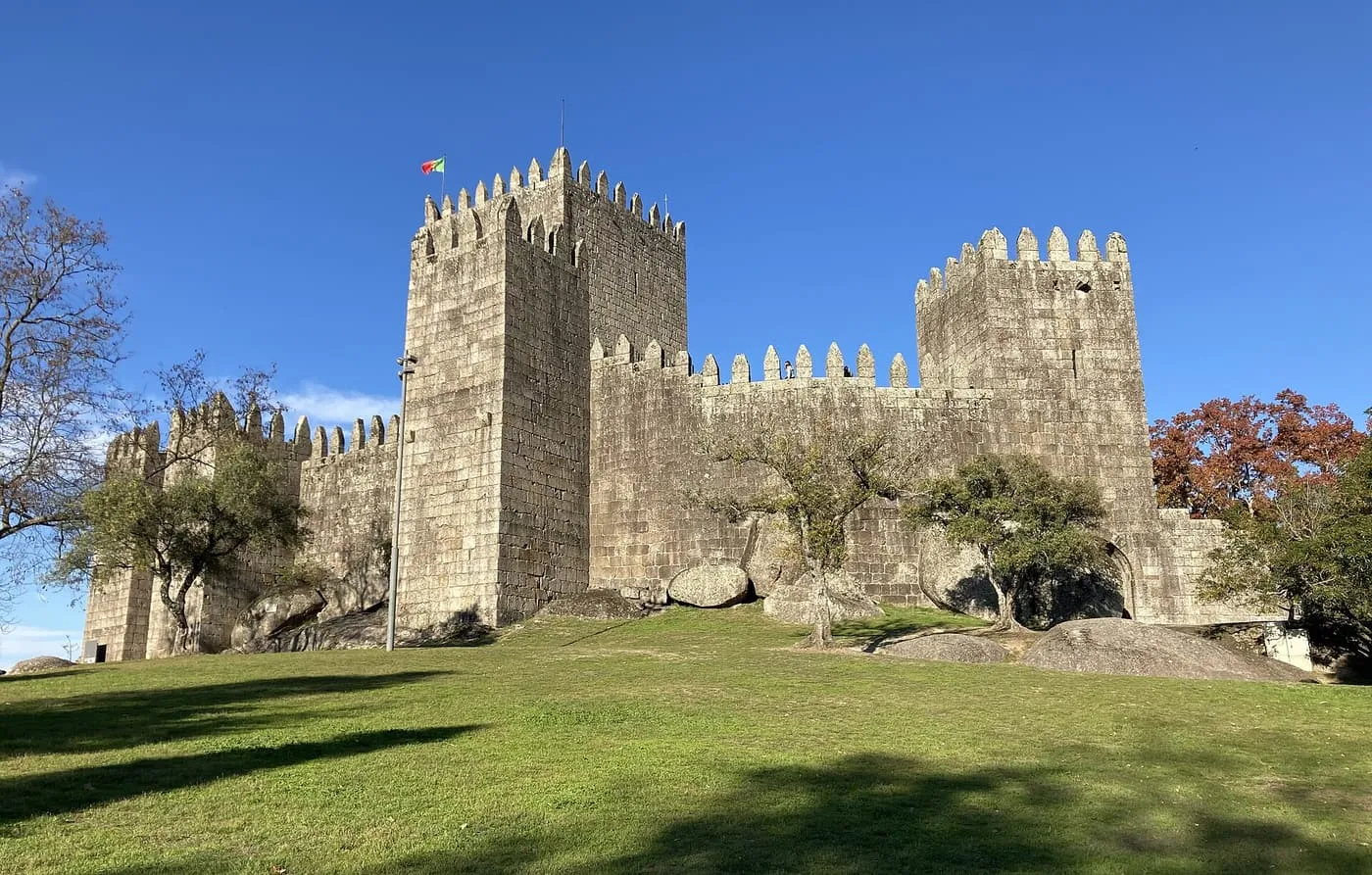 The granite towers and battlements of Guimarães Castle above a grass slope, with a Portuguese flag on the keep